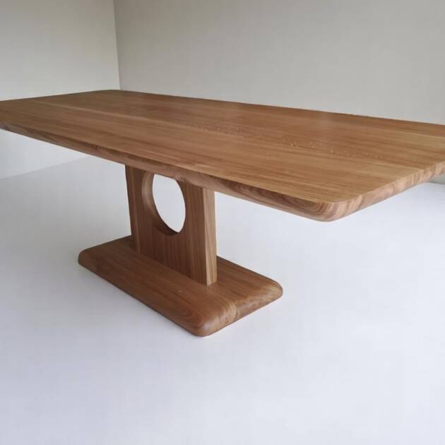 Table de séjour en chêne massif, fabriquée à la main avec un pied circulaire unique, design moderne et bords arrondis, idéale pour un intérieur élégant et contemporain.